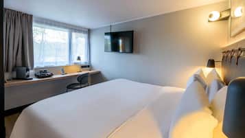 A hotel room with a large bed, a desk with a computer, a chair, a TV mounted on the wall, and a window with curtains.