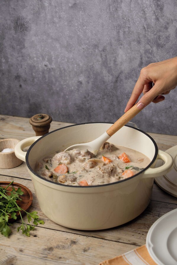 Una olla grande de guiso con una cuchara de madera, rodeada de tazones y un molinillo de pimienta sobre una mesa de madera.