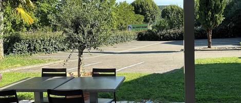 An outdoor patio area with a covered seating arrangement, a small tree, and a parking lot in the background.