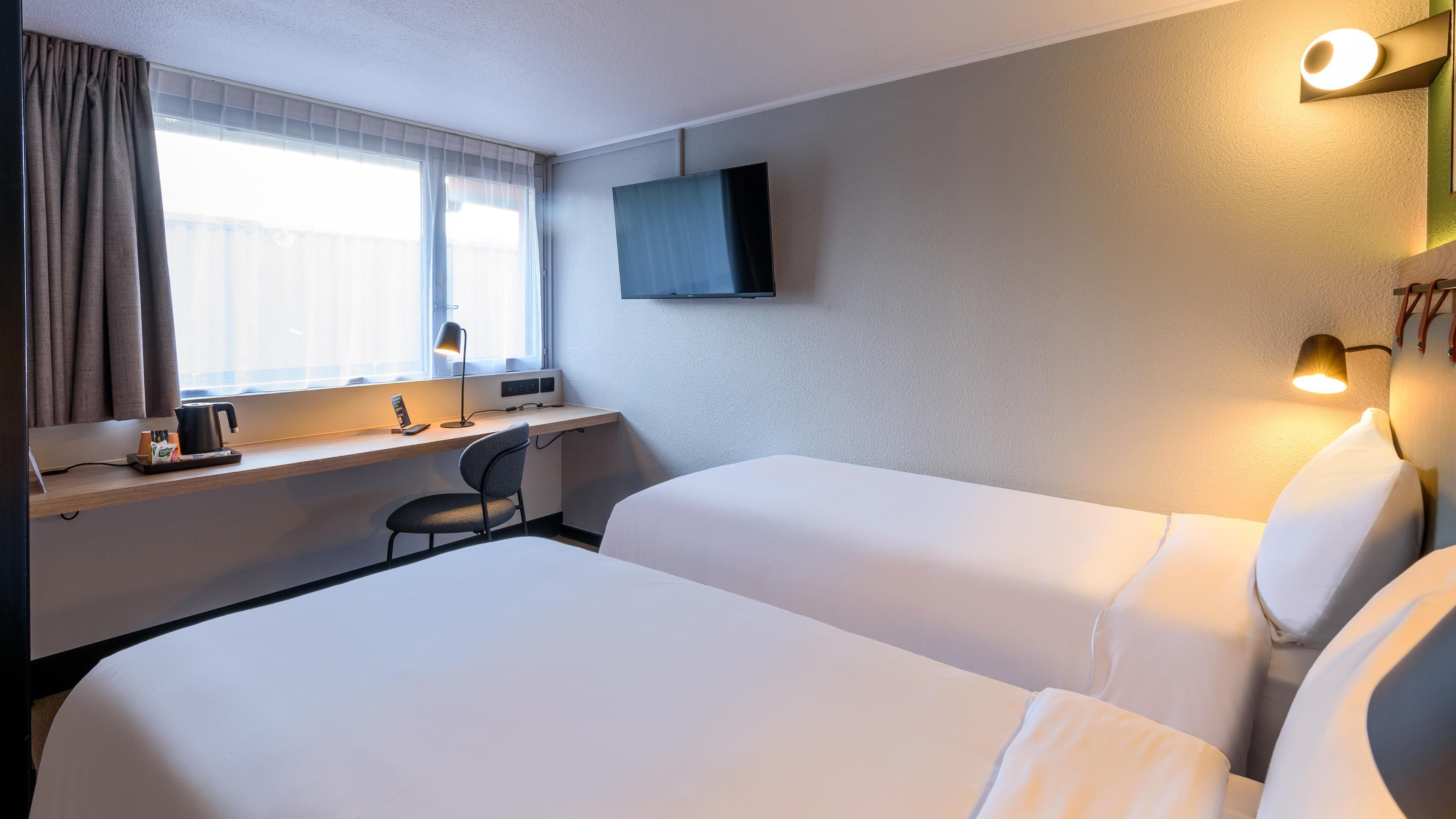 Ein Hotelzimmer mit zwei Betten, einem Schreibtisch mit Stuhl, einem an der Wand befestigten Flachbildfernseher und einem Fenster mit Vorhängen.