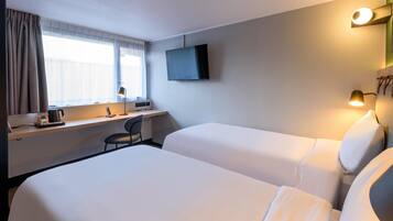 Ein Hotelzimmer mit zwei Betten, einem Schreibtisch mit Stuhl, einem an der Wand befestigten Flachbildfernseher und einem Fenster mit Vorhängen.