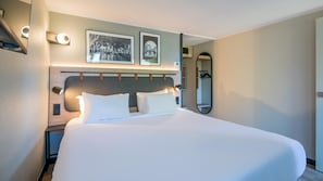 A neatly made bed in a hotel room with a headboard, two framed pictures on the wall, a bedside lamp, and a mirror.