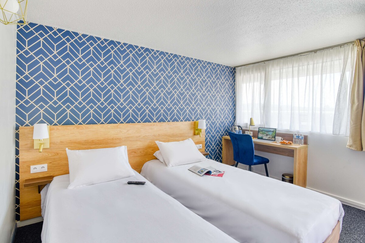 Een hotelkamer met twee bedden, een bureau met een computer en een raam met gordijnen.