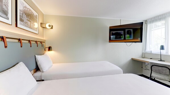 Ein Hotelzimmer mit zwei Betten, einem Schreibtisch mit Computer, einem Fernseher und Wandleuchten.