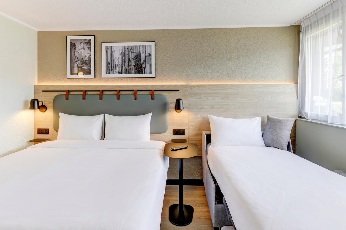 Una habitación de hotel con dos camas, una mesita de noche con un control remoto, lámparas de pared y dos cuadros enmarcados.