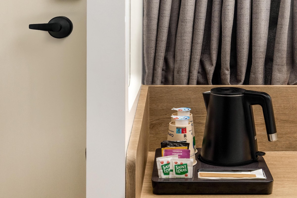 A wooden bedside table with a black electric kettle, a tray with coffee pods, and two Saint Louis coffee packets.