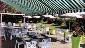 Un patio al aire libre con mesas y sillas de celosía blanca, macetas verdes y toldos a rayas que proporcionan sombra.