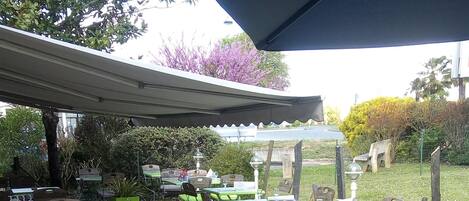 Un espacio al aire libre con mesas y sillas, algunas cubiertas por un toldo, y plantas en macetas.