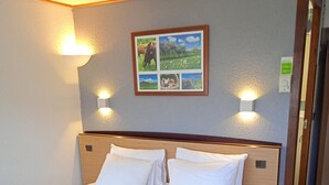 Una cama bien hecha con sábanas blancas, cabecera de madera y una manta azul. Se ve un mesita de noche con un cajón y un mando a distancia encima. La habitación tiene una pared de color claro con un cuadro enmarcado sobre la cama.