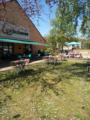 An outdoor seating area with tables and chairs, a building with a sign, and a tree providing shade. - Hotel Campanile Aurillac (Aurillac)