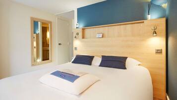 A hotel room with a bed, a wooden headboard, a nightstand, a mirror, and a wall-mounted light fixture.