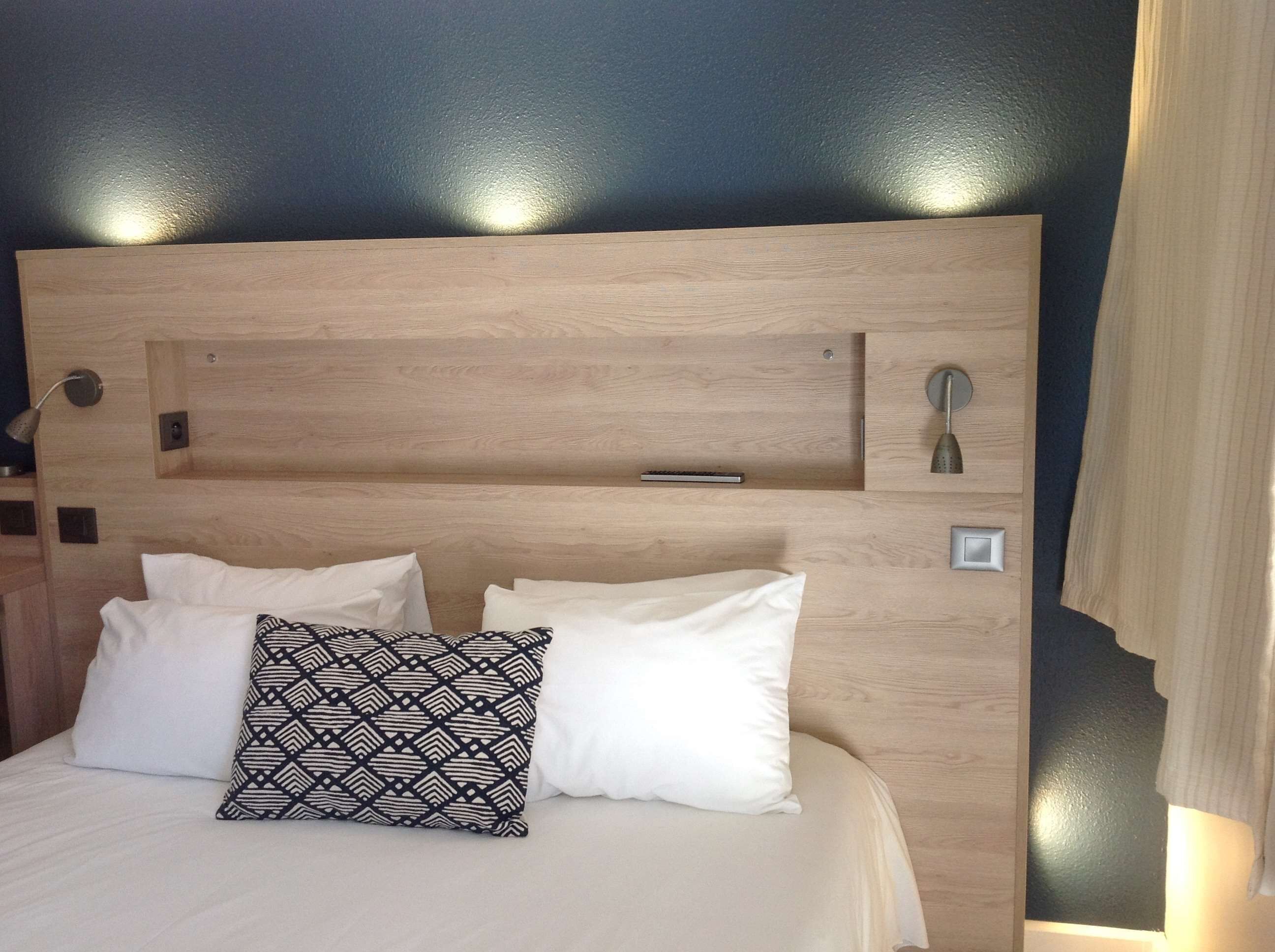 A bed with a wooden headboard, two white pillows, one patterned, and a bedside table with a lamp.