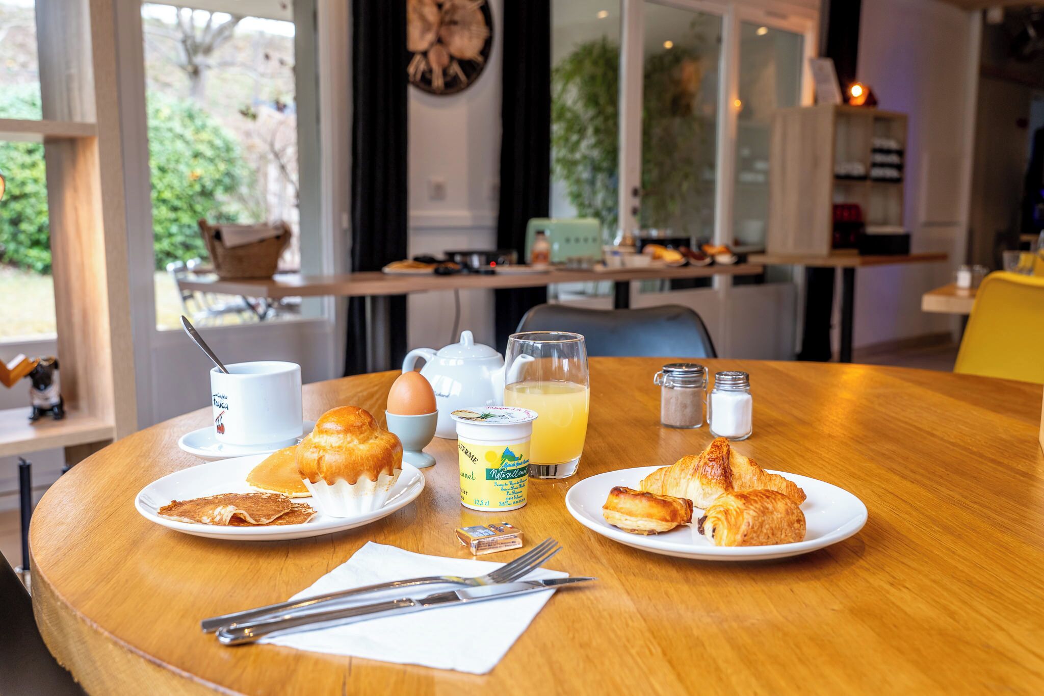 A breakfast table with croissants, a coffee cup, a teapot, a jug of orange juice, and salt and pepper shakers.