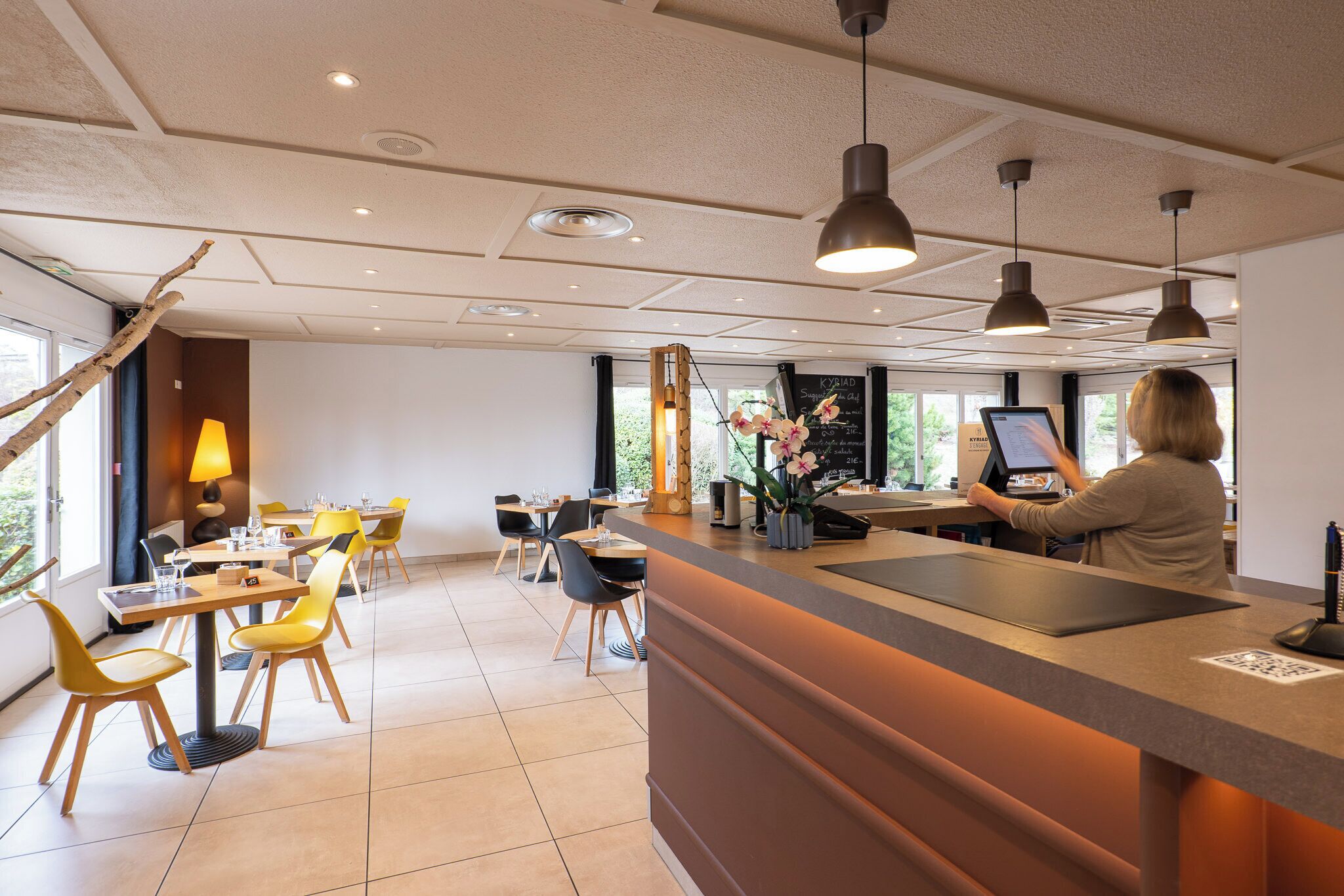 A modern hotel lobby with a reception desk, yellow chairs, and a large window offering a view of the outdoors.