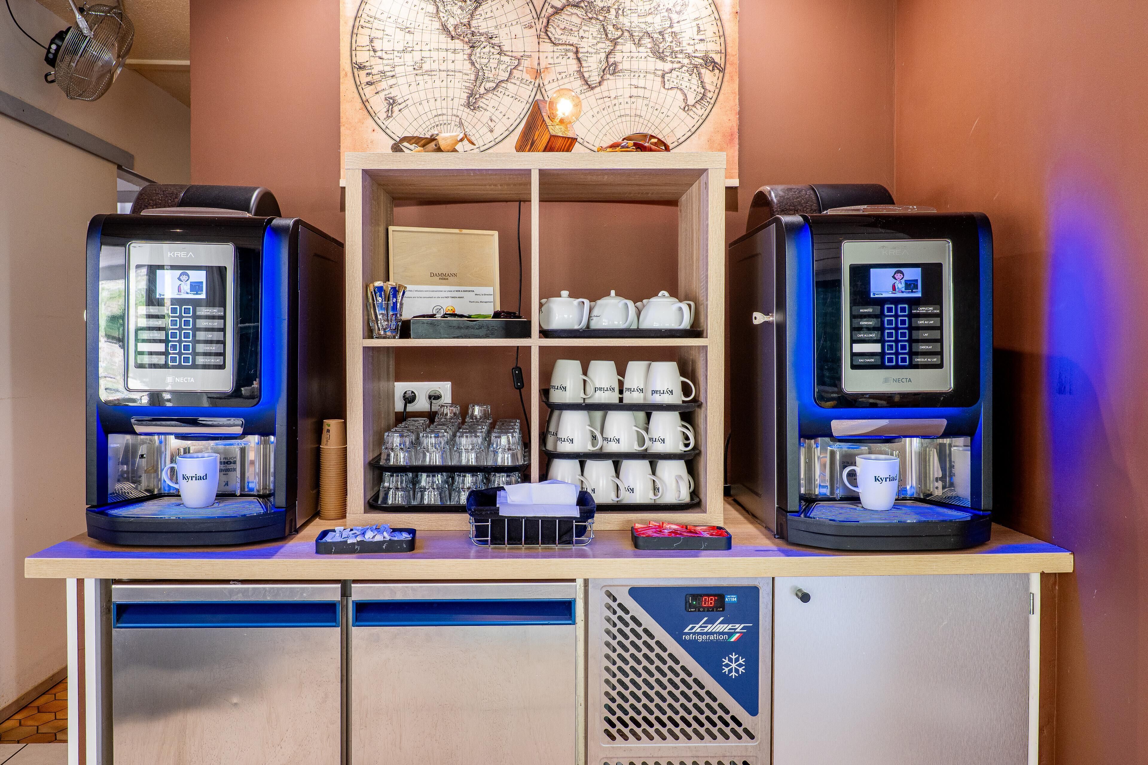 Dos máquinas de café con interfaces digitales, un estante con teteras y tazas, y un refrigerador pequeño debajo.