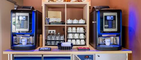 Dos máquinas de café con interfaces digitales, un estante con teteras y tazas, y un refrigerador pequeño debajo.