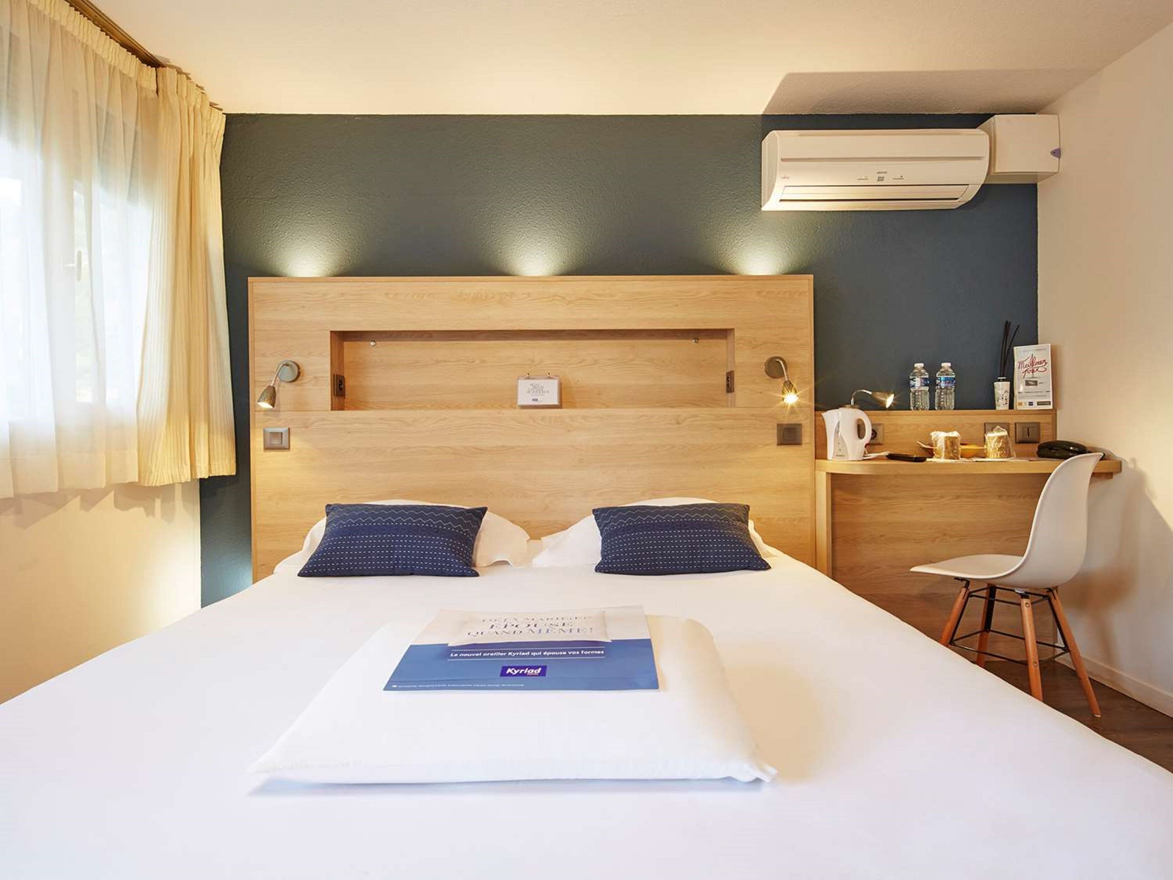 A hotel room with a large bed, two pillows, a bedside table with a kettle and a cup, a chair, and an air conditioning unit on the wall.
