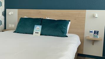 Un lit bien fait, avec des draps blancs et deux coussins turquoise, une tête de lit en bois et une petite étagère sur laquelle se trouvent une bouteille d’eau et une brochure.
