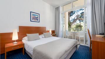 Una habitación de hotel con una cama grande, una mesita de noche de madera con una lámpara, un escritorio con una silla, y un balcón con una mesa y sillas.