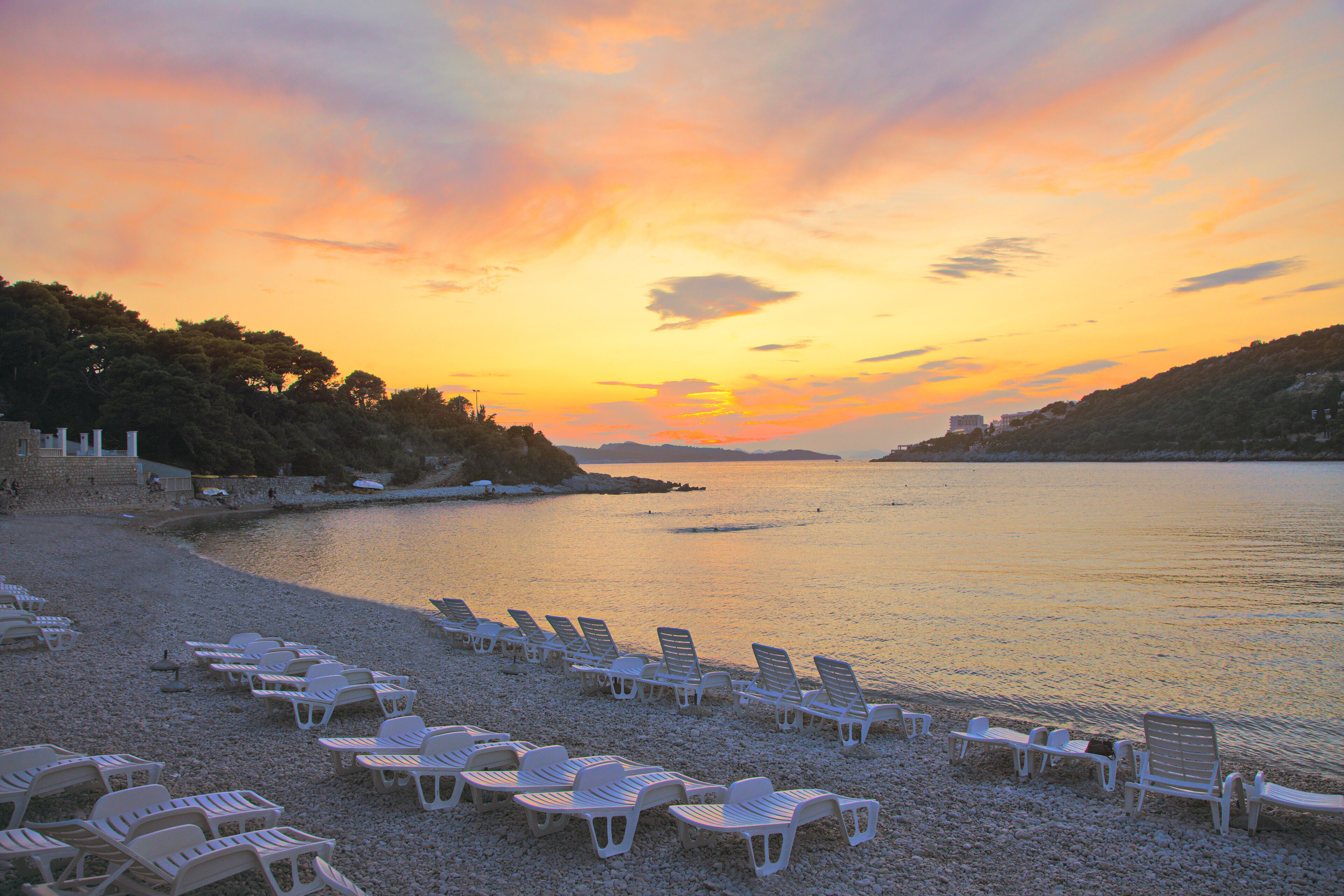 Une plage bordée de rangées de chaises longues blanches, toutes orientées vers la mer, un rivage rocheux et un coucher de soleil en arrière-plan.