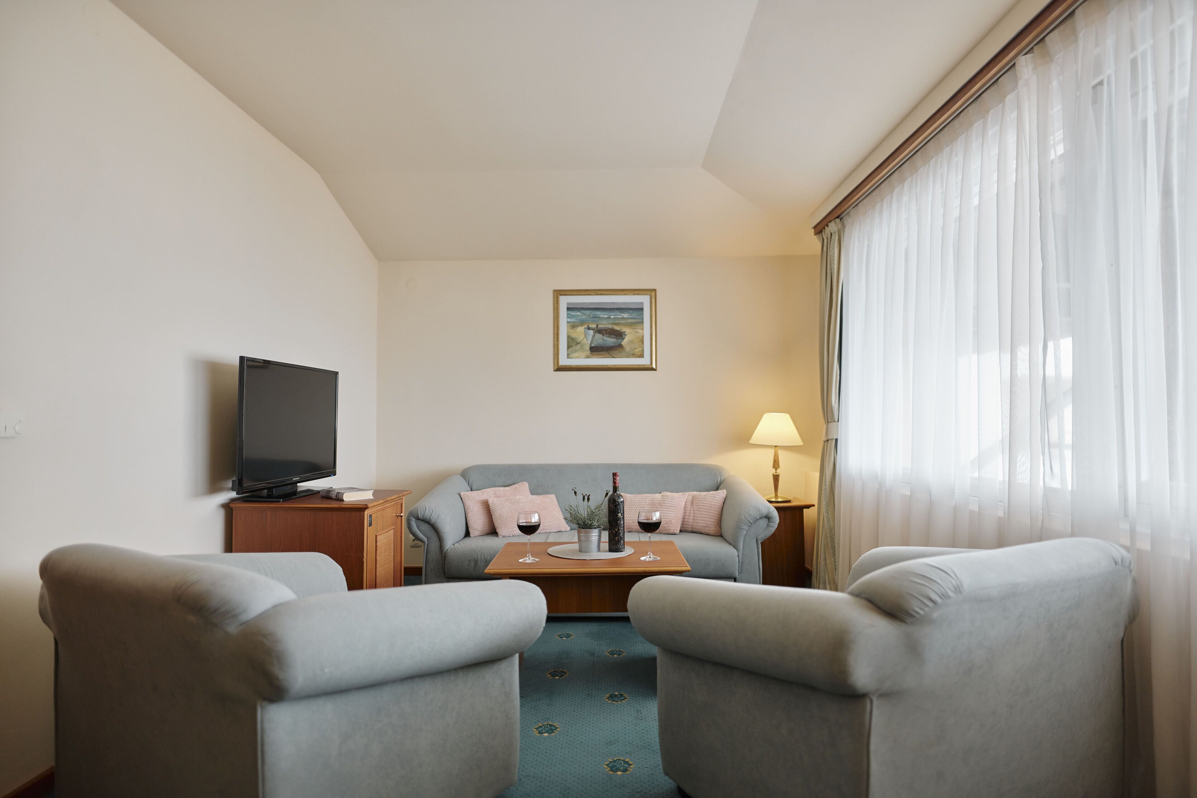 A hotel room with a flat-screen TV, a sofa, two armchairs, a small table with a bottle and glasses, and a painting on the wall.