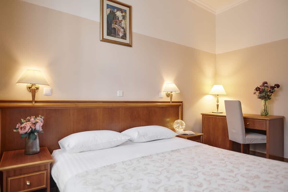A hotel room with a large bed, two bedside lamps, a wooden nightstand with a vase of flowers, a desk with a chair, and a painting on the wall.
