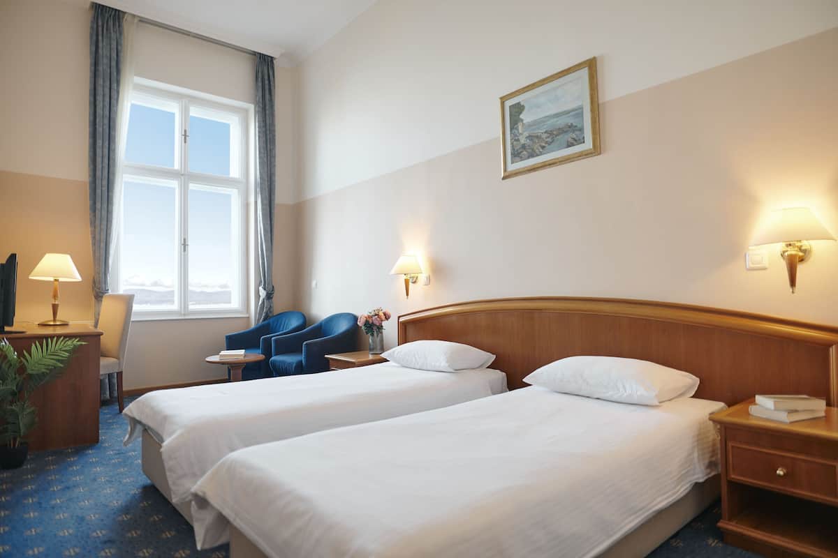 A hotel room with two beds, a desk with a lamp, a chair, a small table, and a window with curtains.