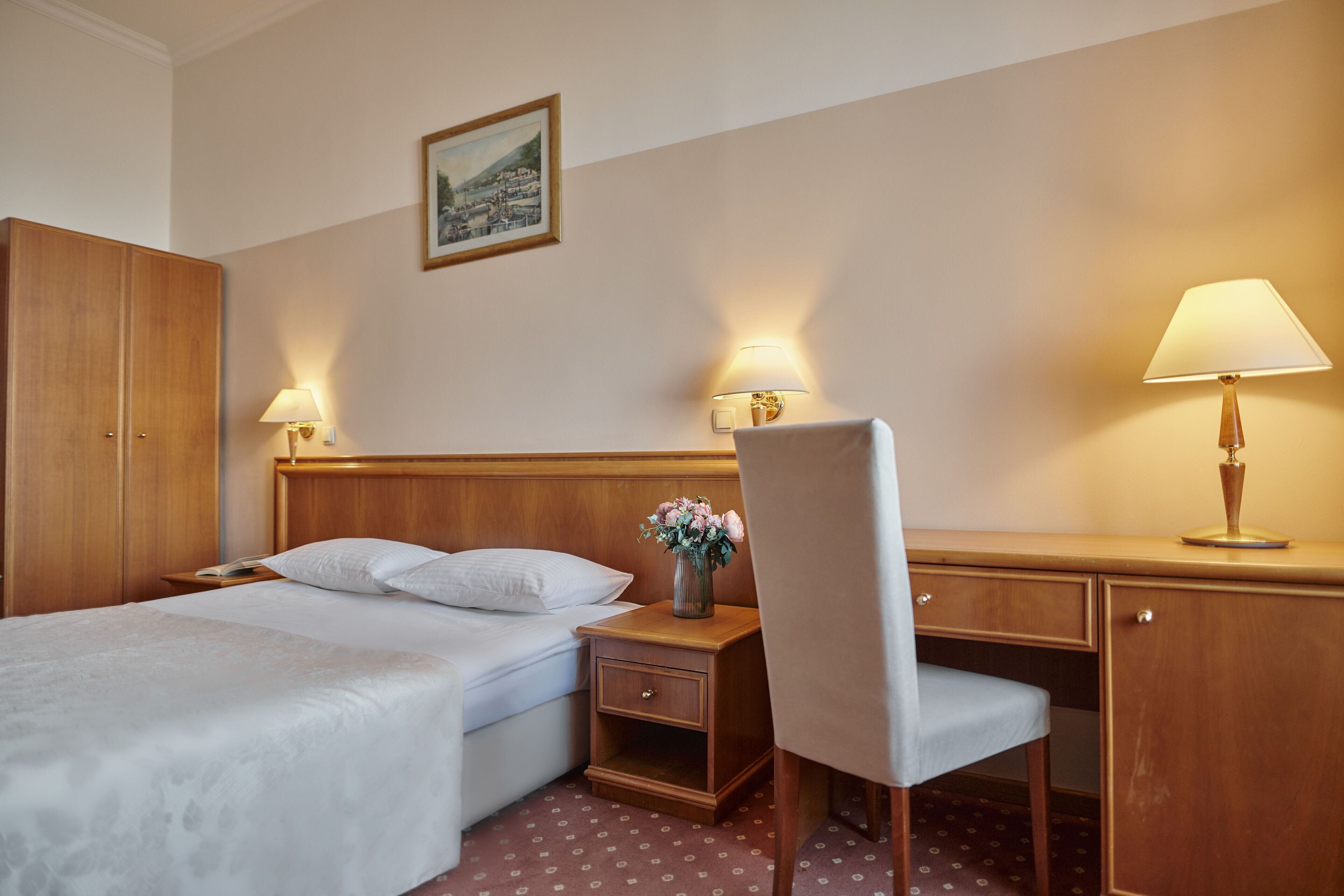 A hotel room with a bed, a desk, a chair, a nightstand, and two lamps.
