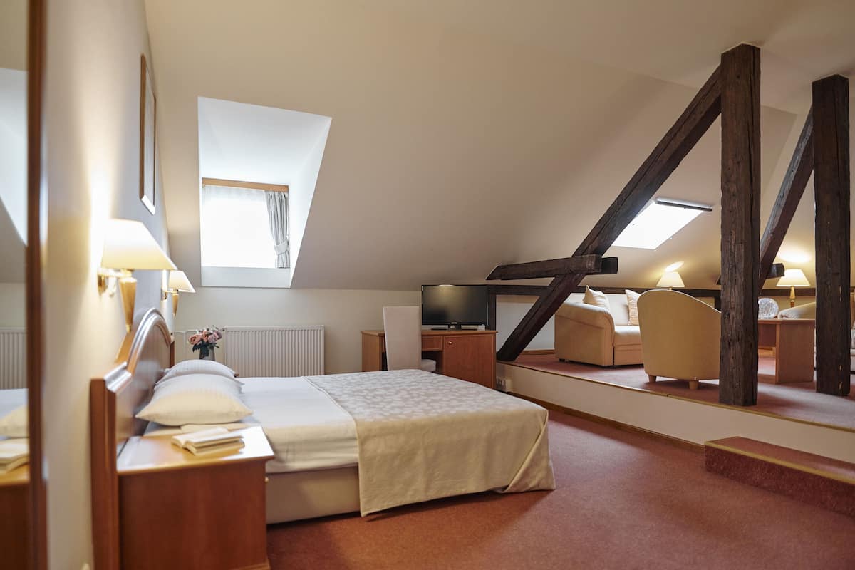 A modern hotel room with a sloped ceiling, a large bed, a desk with a chair, a television, and a large mirror.