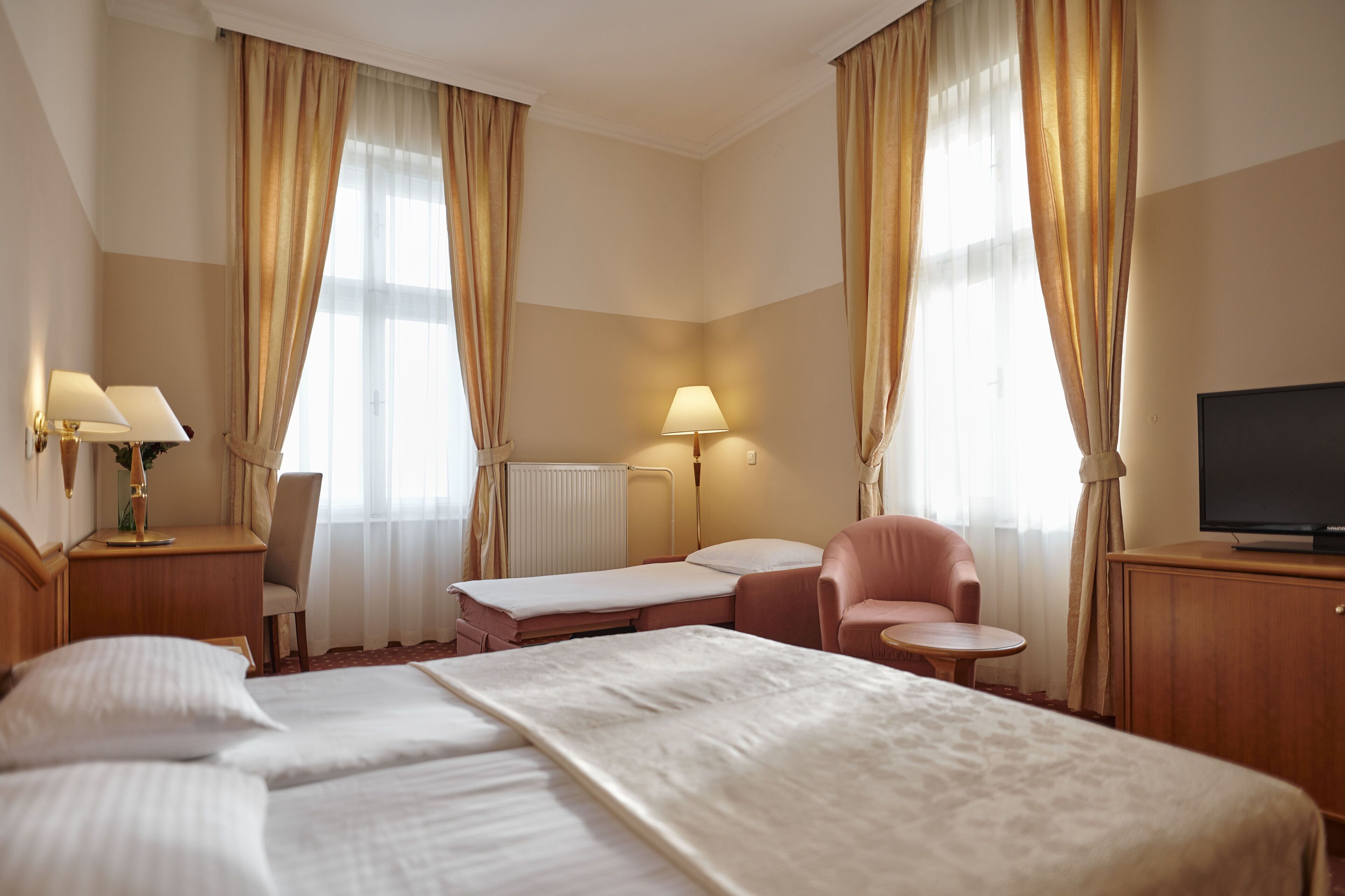Une chambre d’hôtel avec un grand lit, deux tables de chevet avec des lampes, un bureau avec une chaise, une petite table ronde et un téléviseur posé sur un meuble.