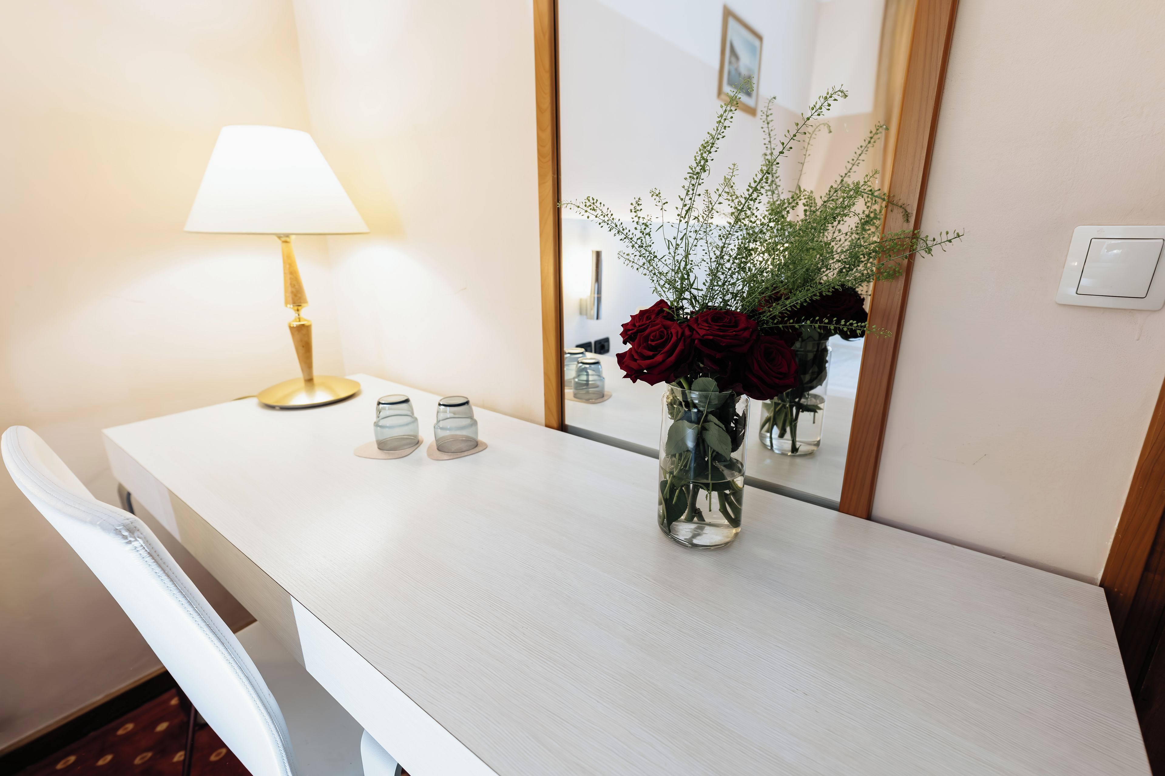 Ein Hotelzimmer mit weißem Schreibtisch, einer Lampe, einer Vase mit roten Rosen und einem Spiegel.