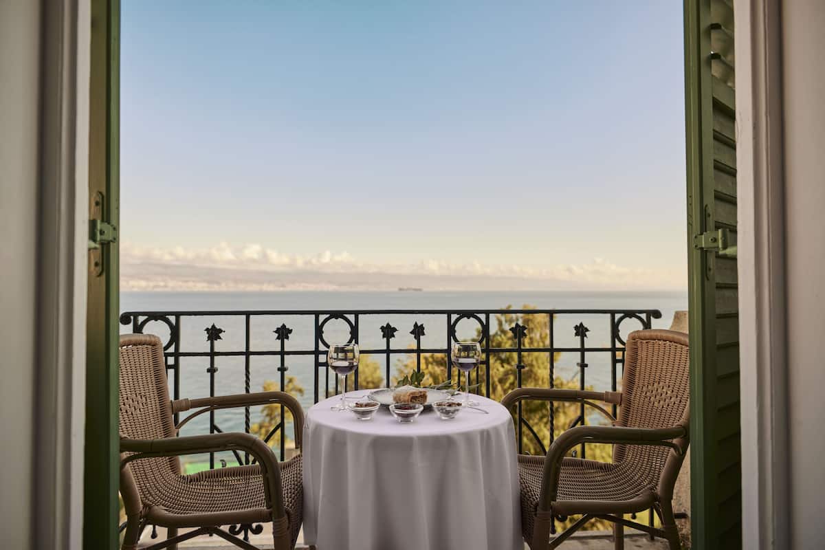 A balcony with wicker chairs and a table set for two, overlooking a scenic view of the sea and mountains.