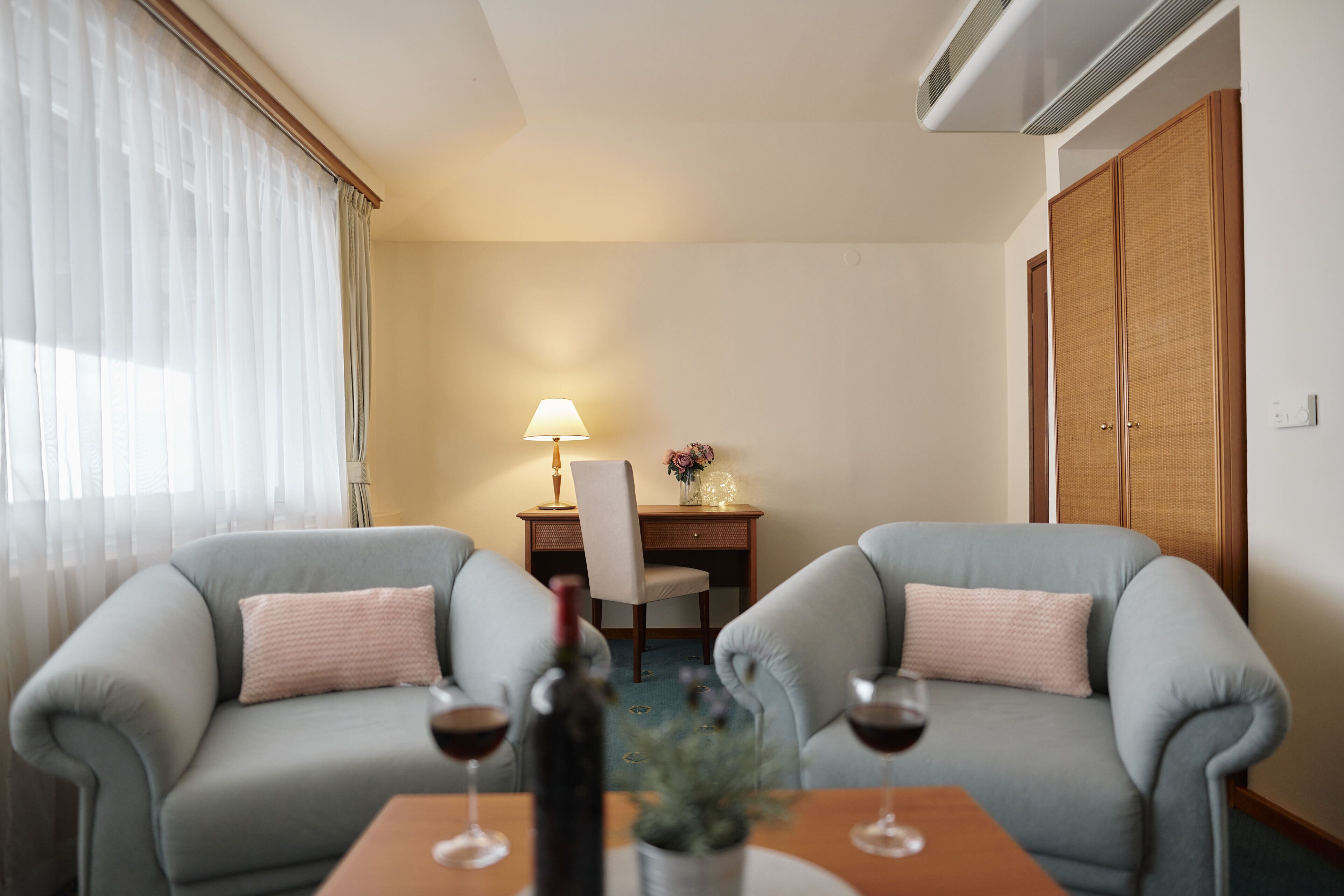 A hotel room with two grey sofas, a wooden table with wine glasses, a lamp, and a desk with a chair.