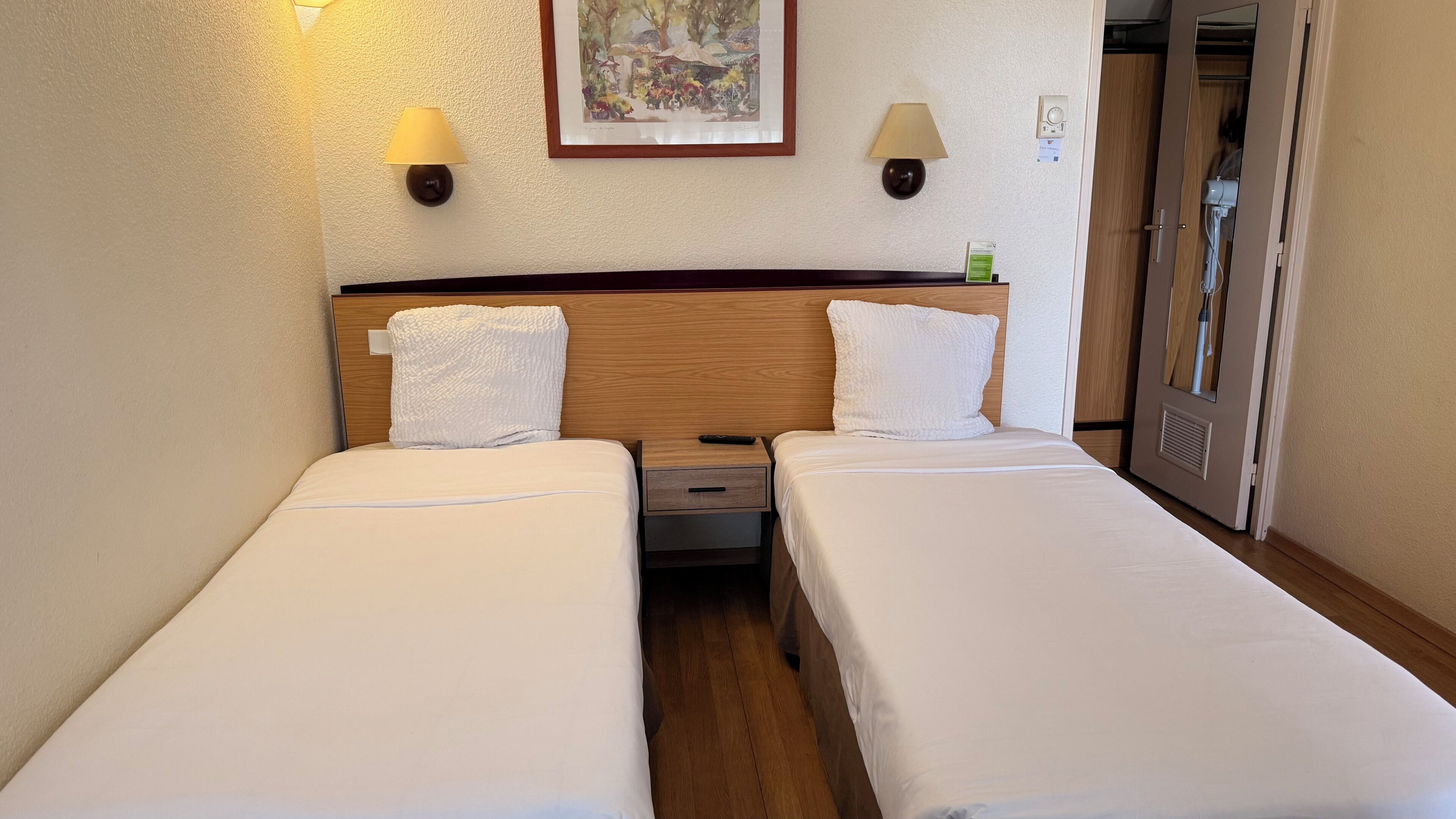 Habitación de hotel con dos camas individuales, cabecera de madera, lámparas de noche, mesitas de noche y un cuadro en la pared.
