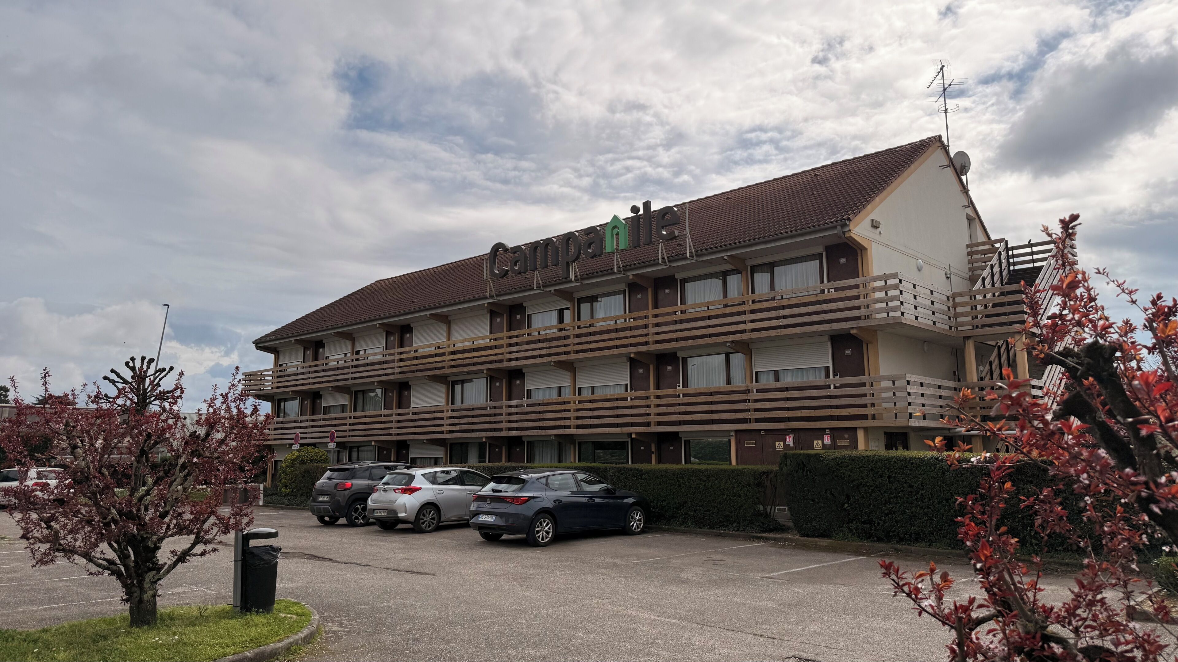 A multi-story hotel with a flat roof, balconies, and a parking lot with several cars.