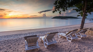 A beach with three white lounge chairs under a tree, a calm sea, and a sunset. - Nora Beach Resort and Spa (Koh Samui)