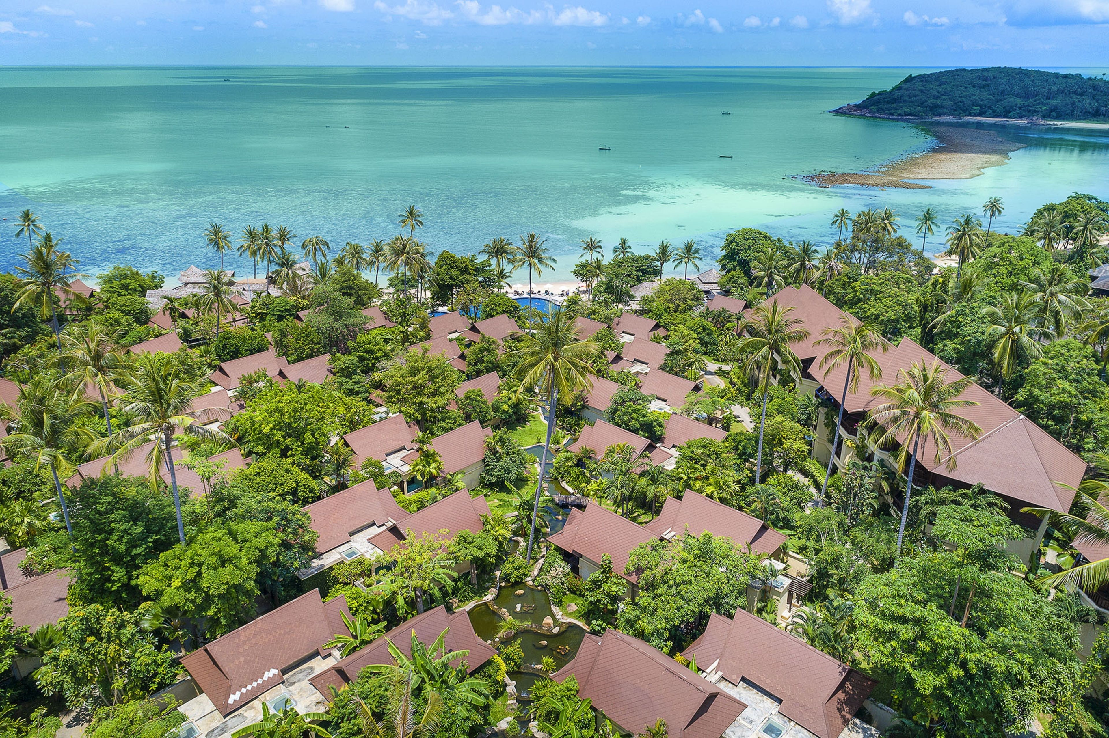 An aerial view of a resort with multiple bungalow-style buildings, a central pool, and a beach area.