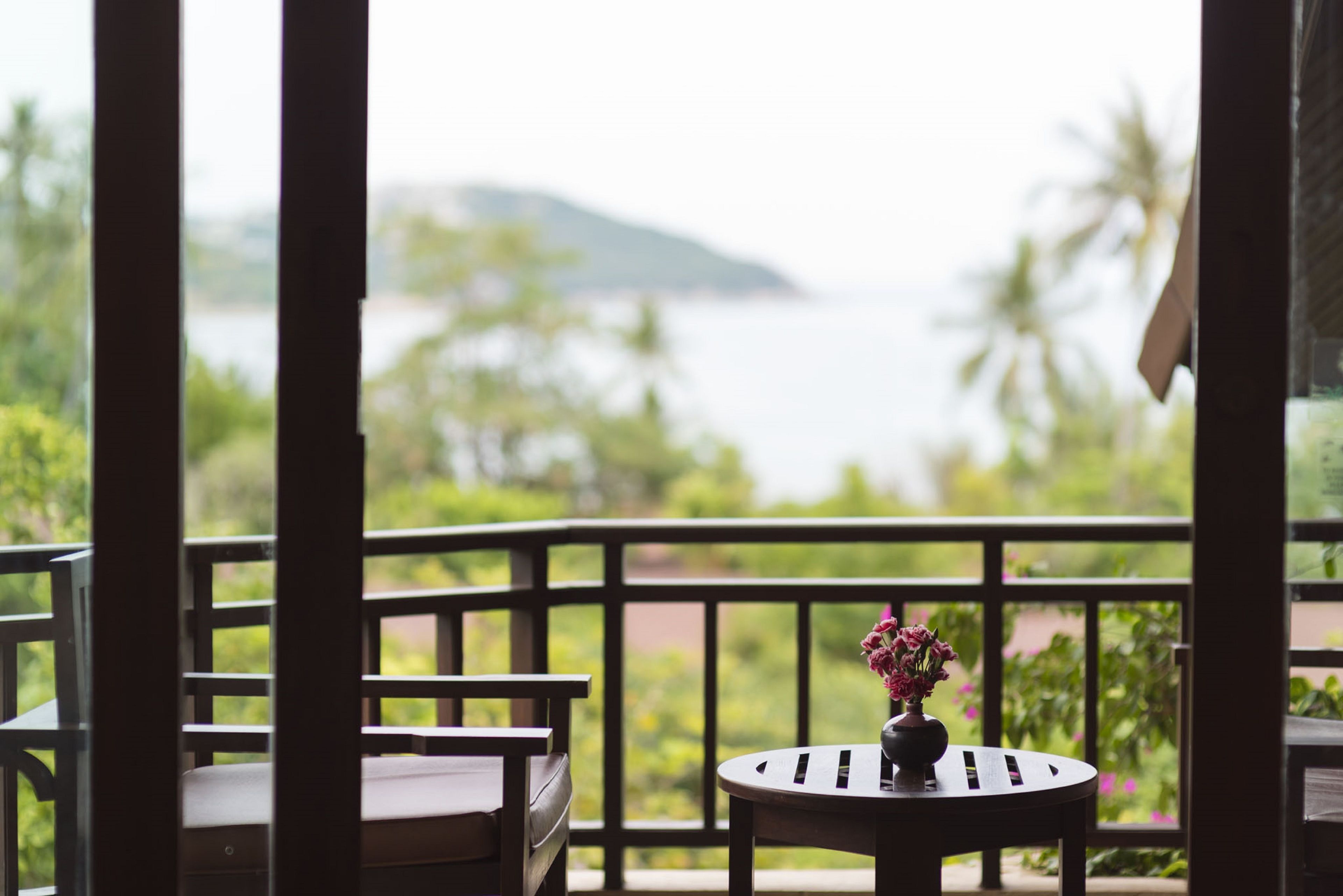 A balcony with a wooden railing, a round table with a vase of flowers, and a view of a body of water and greenery.