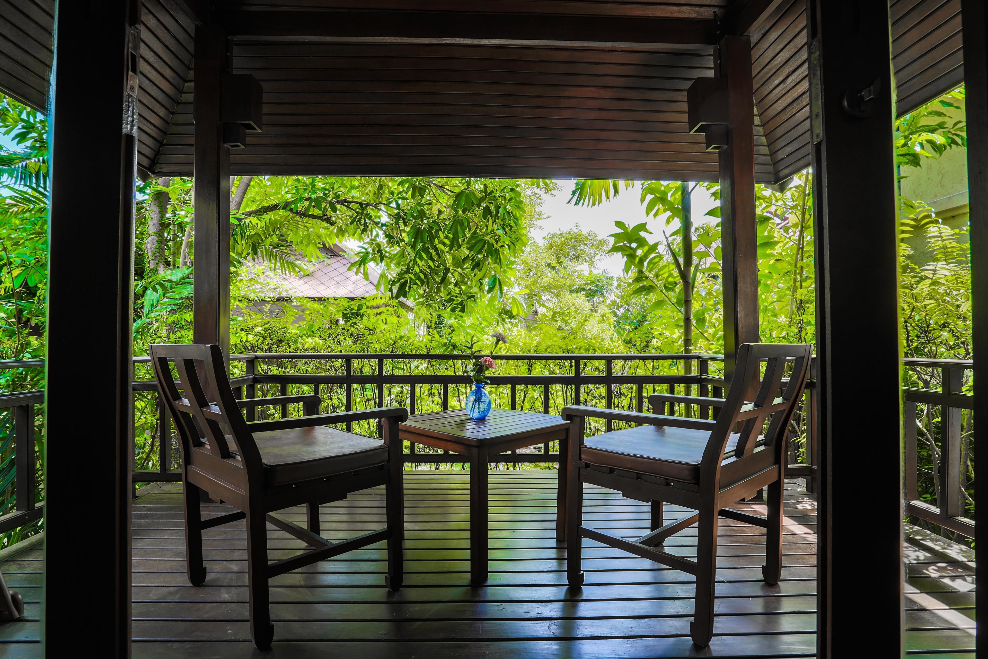 A wooden deck with two chairs and a small table, surrounded by a railing, overlooking a lush garden.