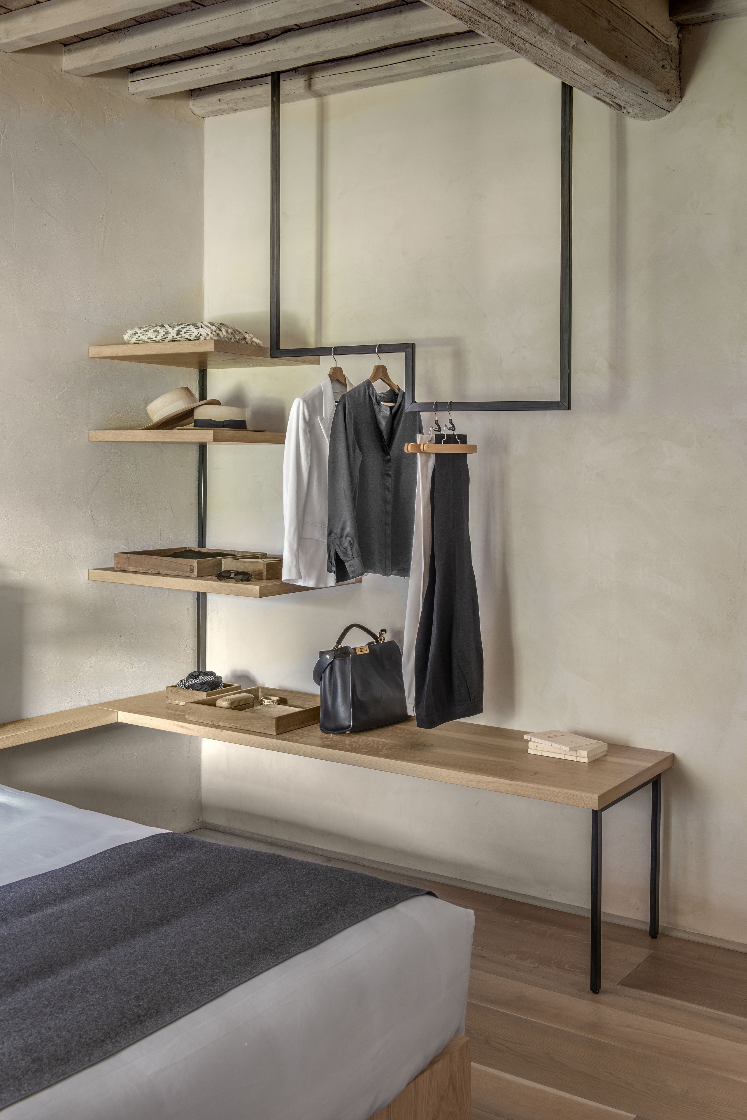 Une chambre à coucher avec un lit, un bureau en bois, des étagères avec des vêtements et des sacs, et un miroir au mur.
