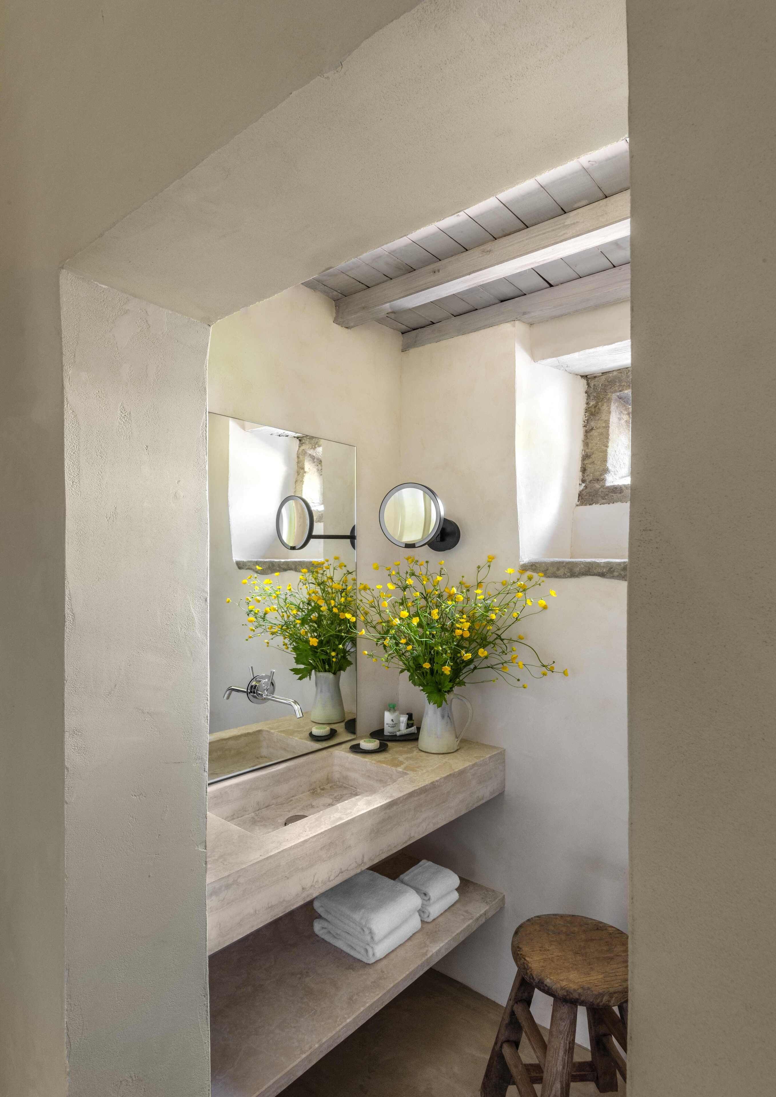 A bathroom with a large mirror, two sinks, a wooden stool, and a vase with yellow flowers.