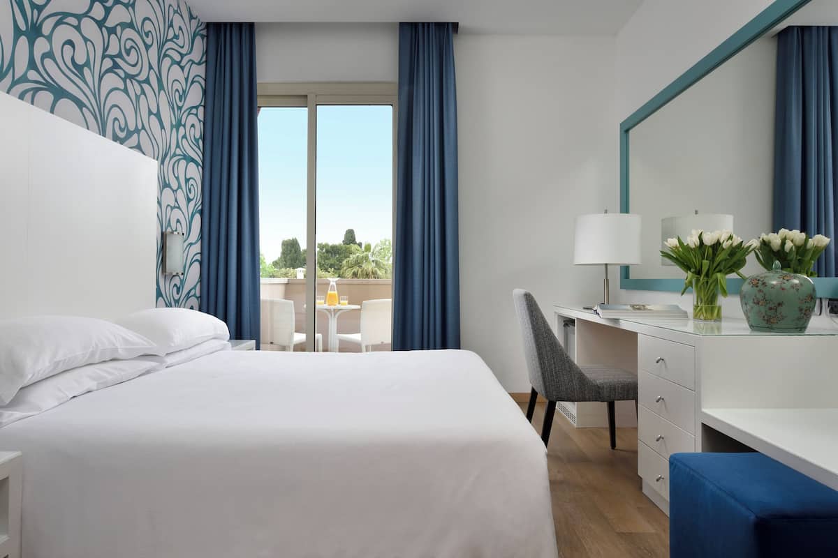 Habitación de hotel con una cama grande, un escritorio con silla, una mesita de noche con lámpara y vistas al exterior.