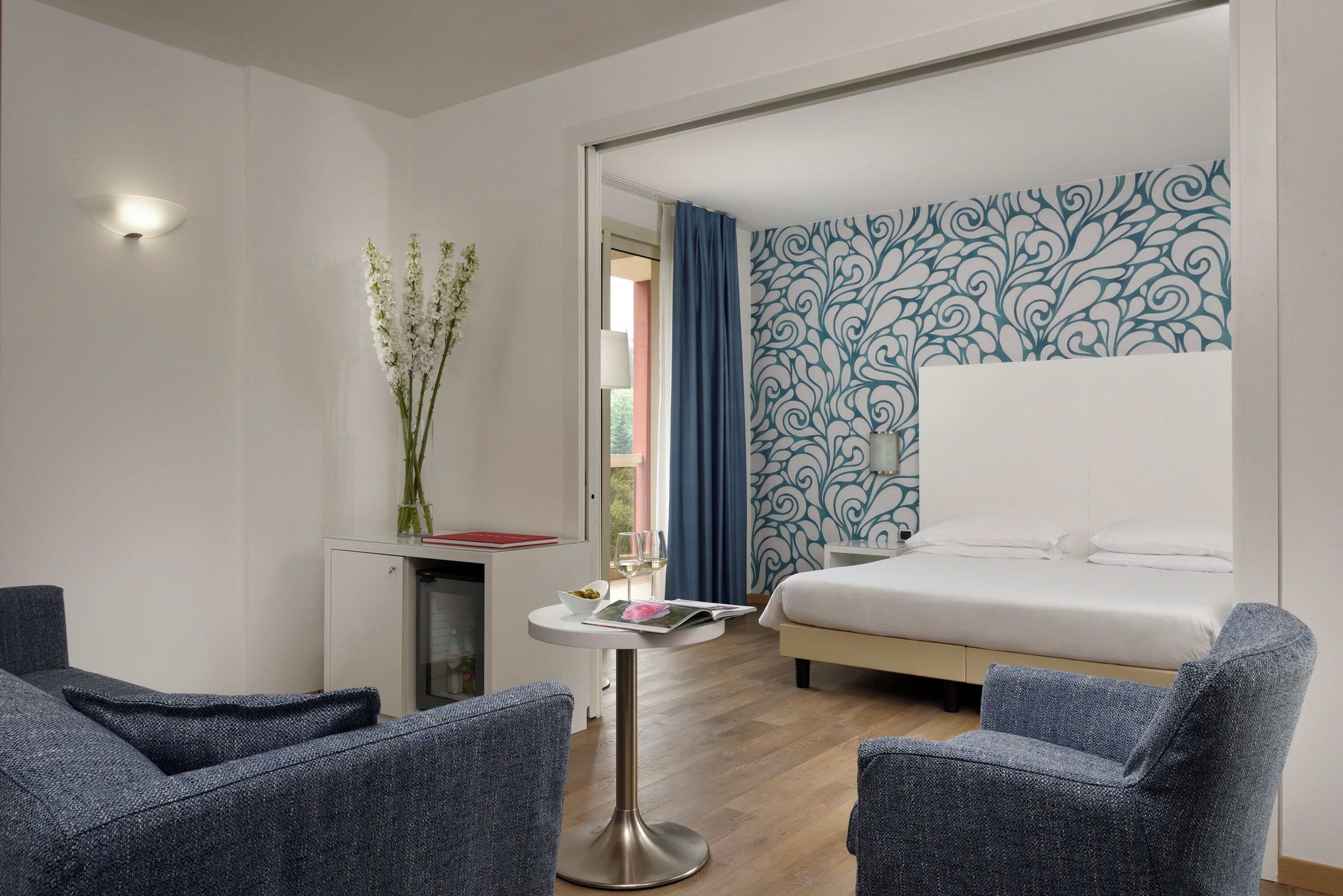 A modern hotel room with a bed, two armchairs, a small table with a vase of flowers, and a decorative wall.