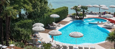 A large outdoor swimming pool with a view of the sea, surrounded by lounge chairs and umbrellas.