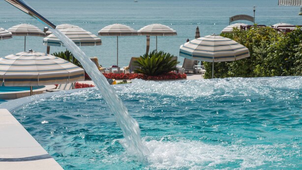 Una piscina con agua que se desliza por el borde, rodeada de sillas de descanso y sombrillas.