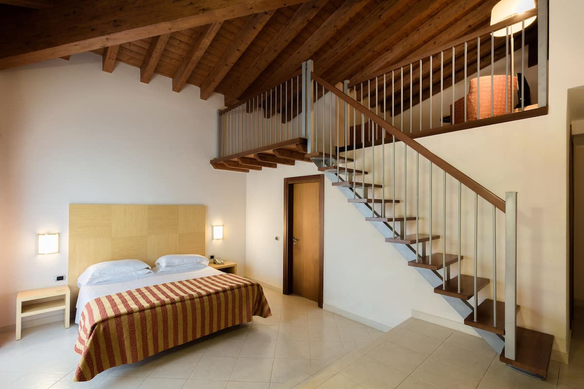 Un dormitorio con cabecera de madera, cama con colcha a rayas, mesita de noche, lámpara de escritorio y una escalera con barandilla.