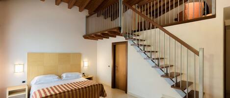 Un dormitorio con cabecera de madera, cama con colcha a rayas, mesita de noche, lámpara de escritorio y una escalera con barandilla.