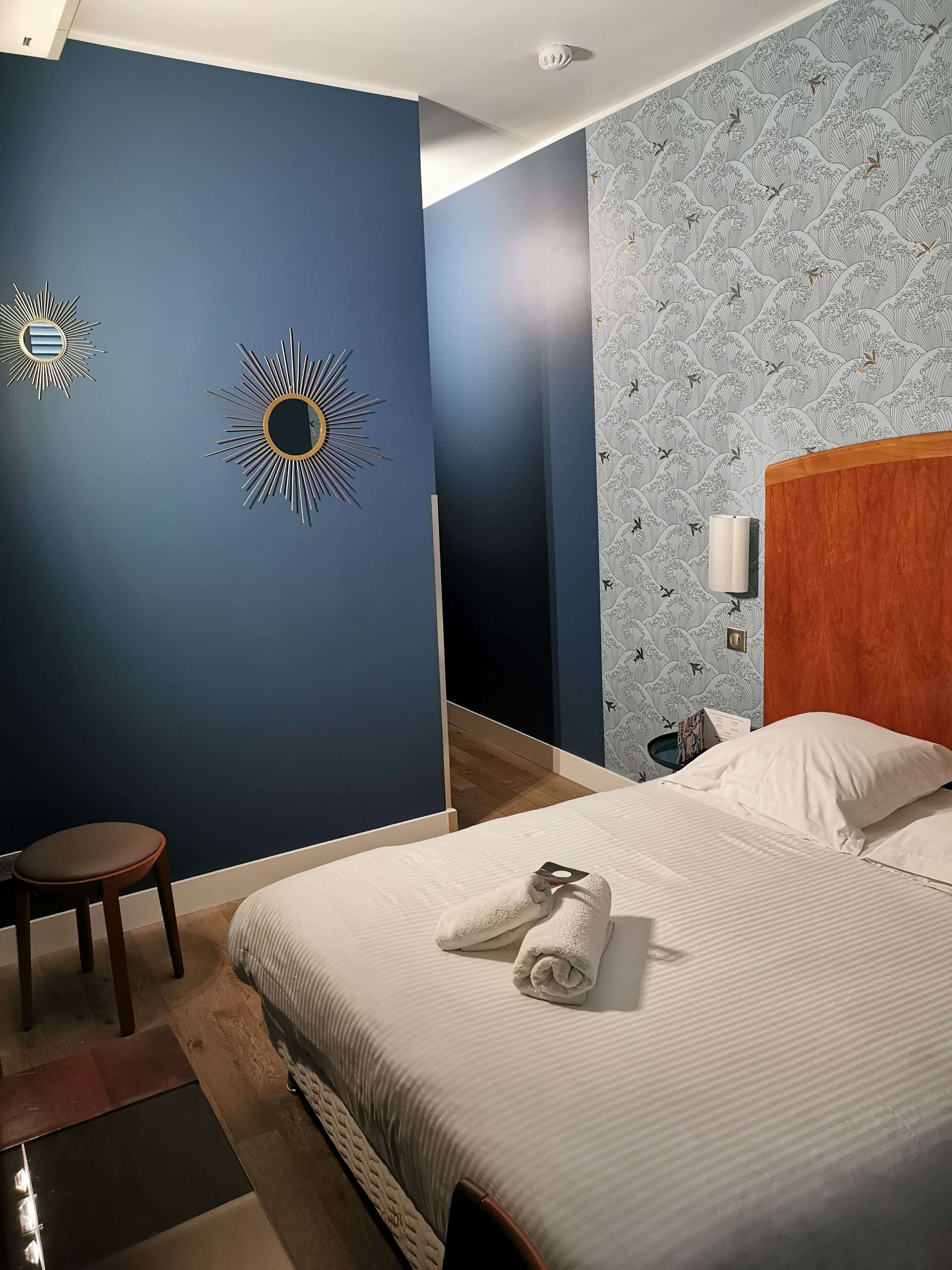 A hotel room with a bed, a nightstand, a wall-mounted lamp, and a chair.