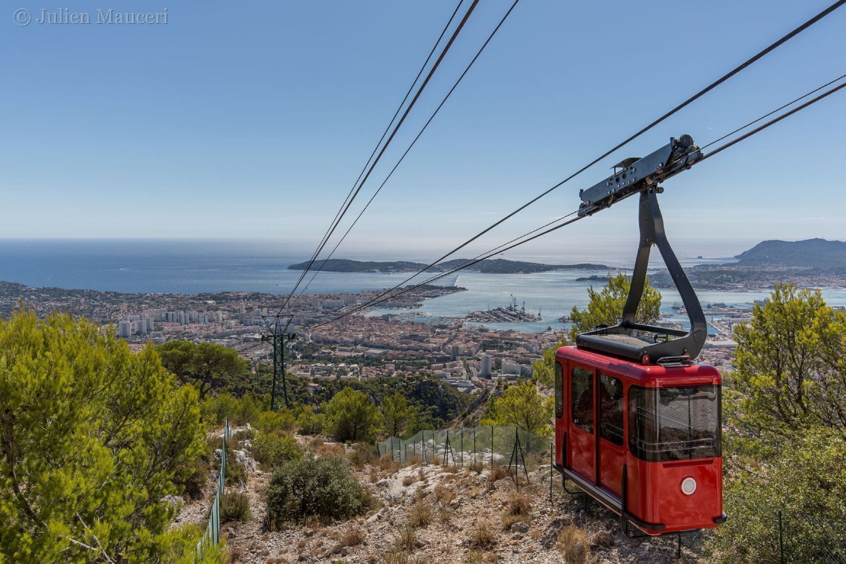 A cable car system with a red gondola, a clear view of a coastal cityscape, and a body of water in the distance.