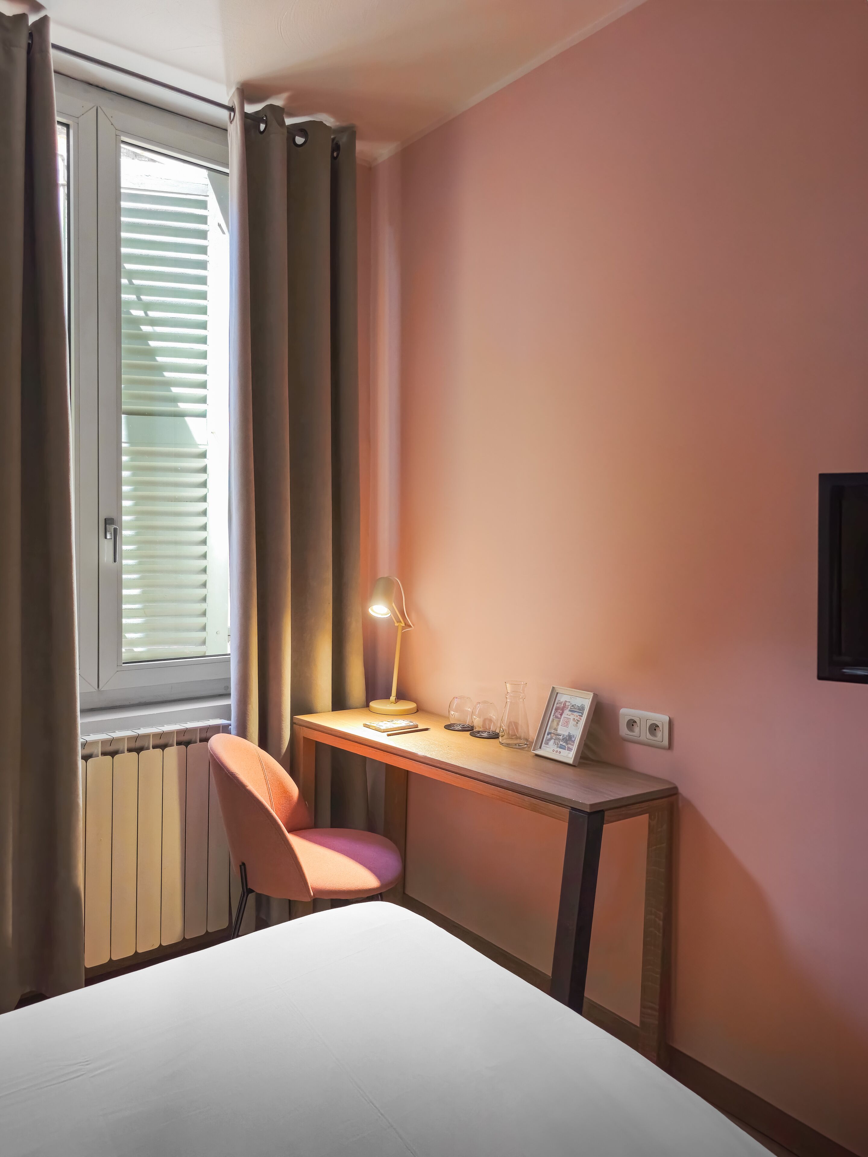 Una camera d'albergo con un letto, una scrivania con una lampada, una sedia e una finestra con tende.